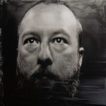 Black-and-white close-up portrait of a bald person with a full beard, gazing slightly upward against a dark, textured background.