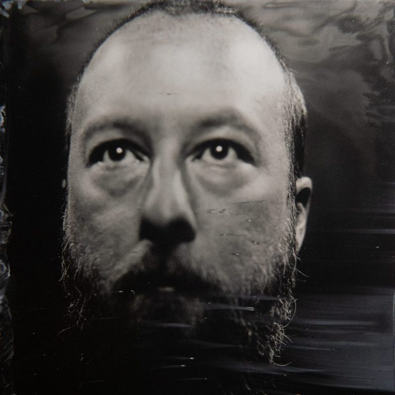 Black-and-white close-up portrait of a bald person with a full beard, gazing slightly upward against a dark, textured background.