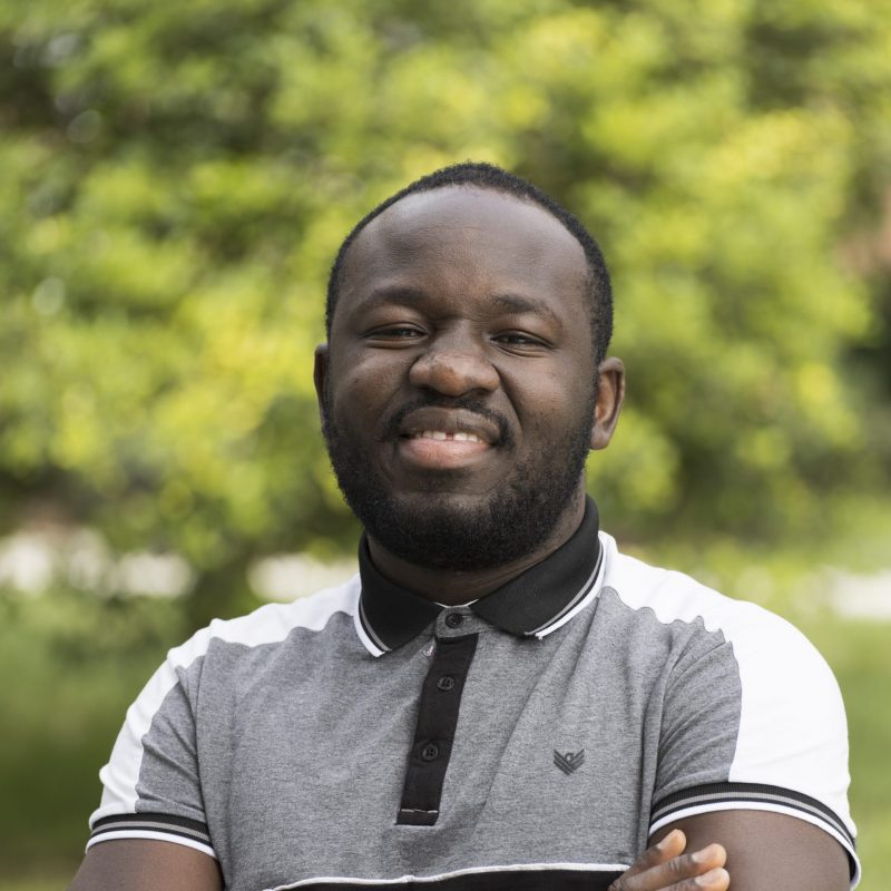 Portrait of a smiling man with a short beard standing outdoors with his arms crossed, wearing a gray-and-black polo shirt, with a softly blurred green background.