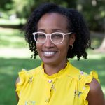 Outdoor portrait of a smiling woman with short curly hair streaked with gray, wearing light-colored glasses and small hoop earrings. She is dressed in a bright yellow ruffled blouse with a subtle floral pattern, standing against a softly blurred green park background.