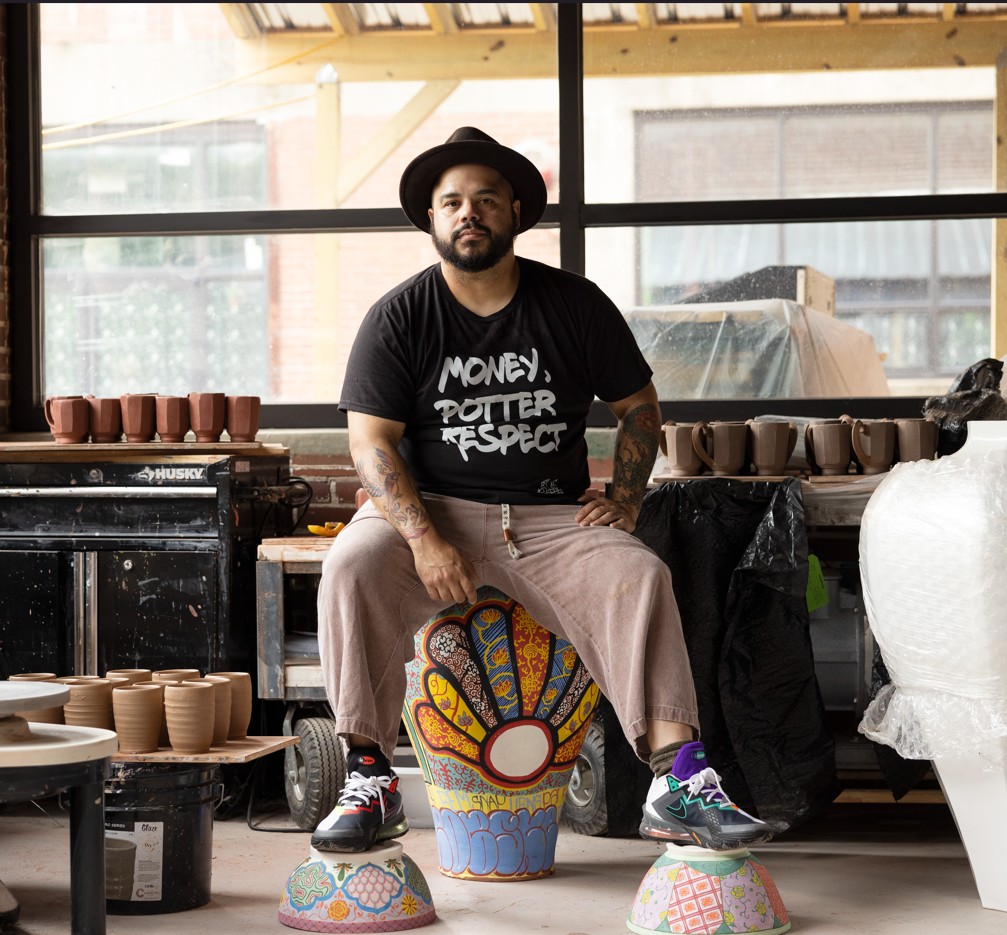 Portrait of a ceramic artist seated in a studio, wearing a black hat, graphic T-shirt, and light pants while resting on painted pottery pieces. Shelves and worktables around him hold rows of clay mugs and vessels, emphasizing an active studio environment.