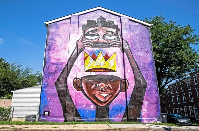 Bold mural on the side of a house showing a stylized Black figure holding up a face or mask with a yellow crown between two faces. Painted in purple, pink, black, and gold tones, the mural fills the entire wall and stands out against a bright blue sky.