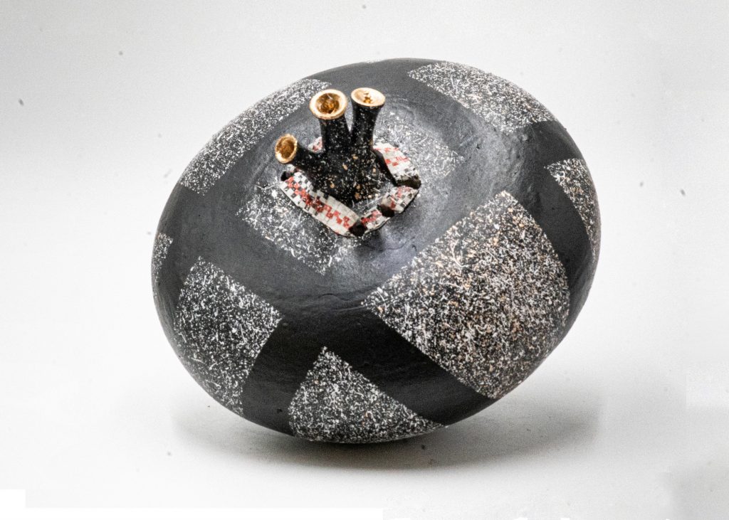 Oval ceramic vessel with a textured black-and-silver surface marked by wide dark bands that wrap diagonally across the form. Three short tubular spouts with copper-colored interiors rise from the top, set on a small square base with patterned detail.