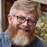 Close-up headshot of a smiling man with blond hair, black rectangular glasses, and a full reddish-blond beard, wearing a blue plaid shirt indoors.