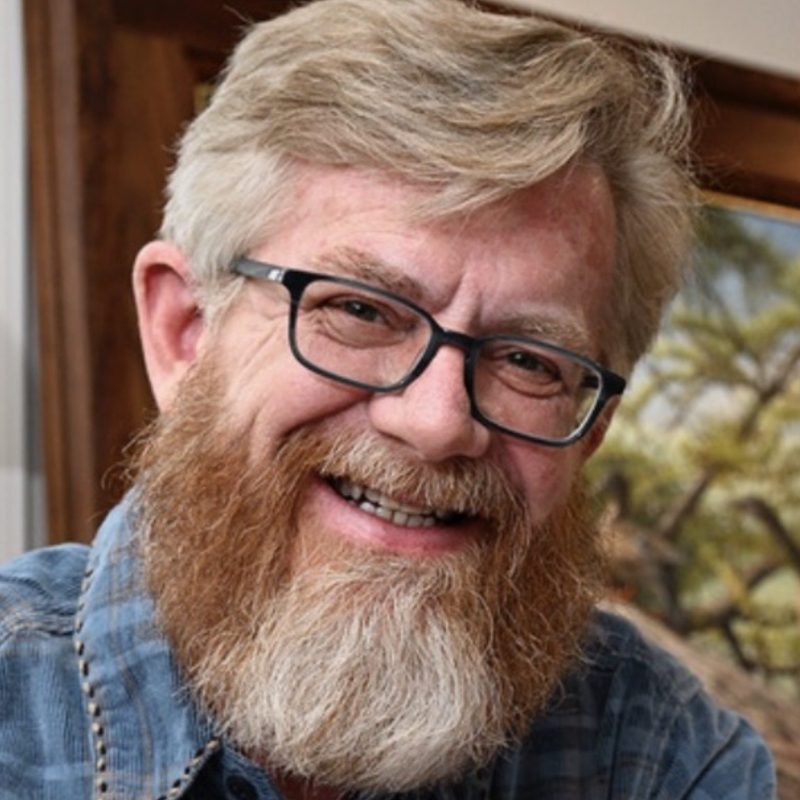 Close-up headshot of a smiling man with blond hair, black rectangular glasses, and a full reddish-blond beard, wearing a blue plaid shirt indoors.