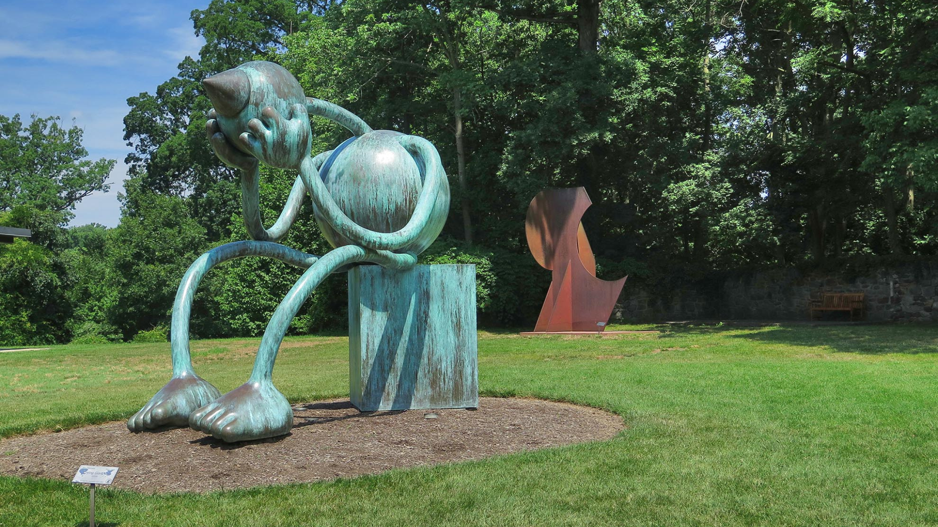 Large outdoor sculpture of a seated, cartoon-like figure with a bulbous head and round body, bent forward with its face resting in one hand as if crying or grieving. The green-patinated metal piece stands on a grassy lawn bordered by tall trees, with another abstract rust-colored sculpture visible in the background.