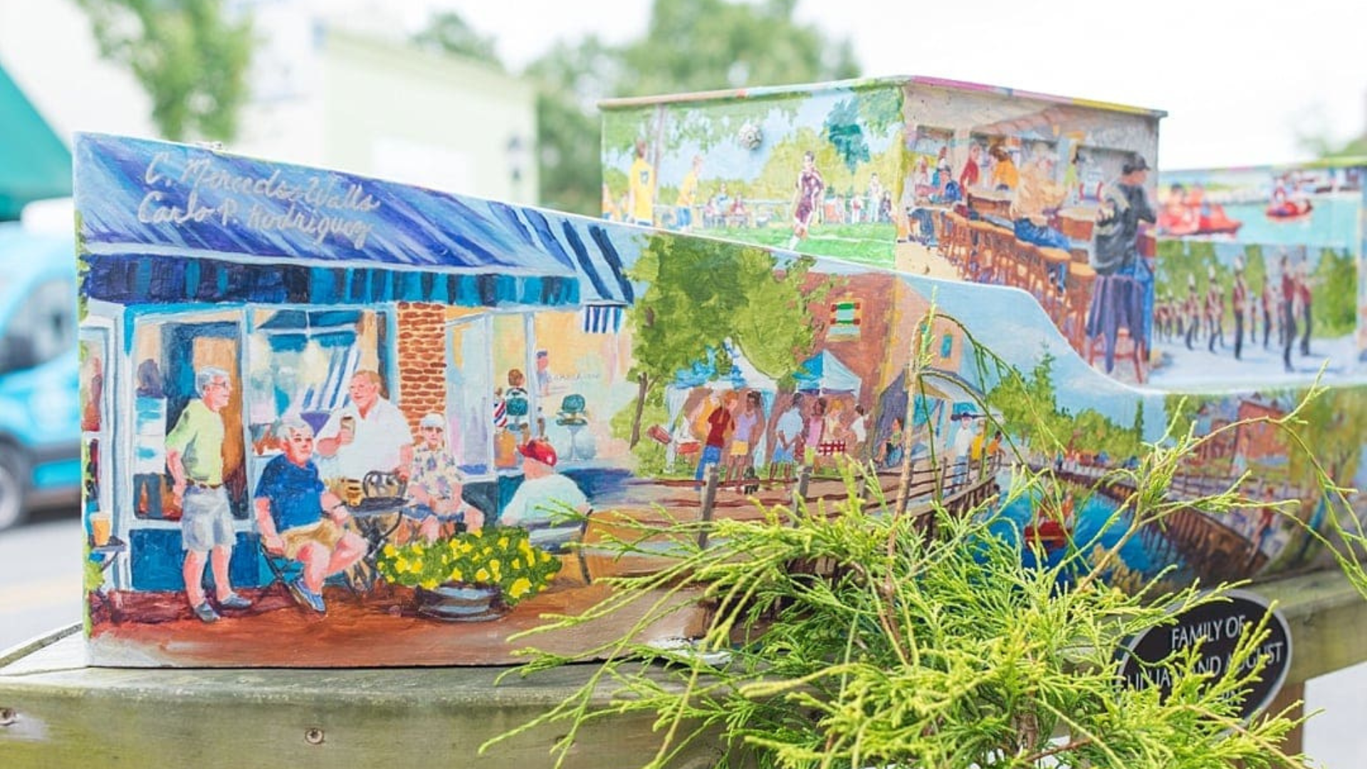 Painted utility box covered with colorful scenes of community life, including people gathered outside shops, dining together, and enjoying a waterfront area. The artwork sits outdoors beside greenery, with the mural wrapping around the box in a bright, small-town montage.