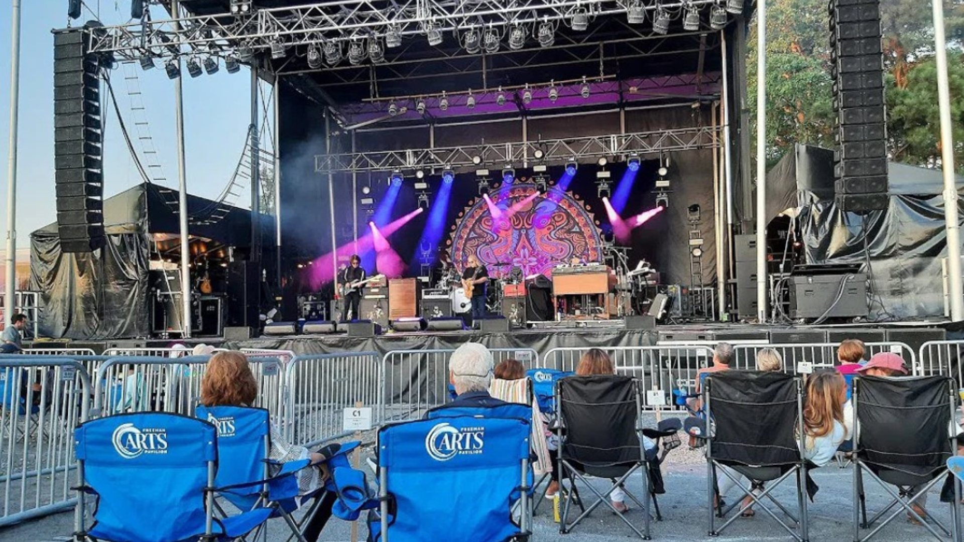 Outdoor concert scene with a band performing on a large festival stage lit in purple and blue. Audience members sit in folding chairs behind metal barriers, facing the stage and sound equipment at what appears to be an evening live music event.
