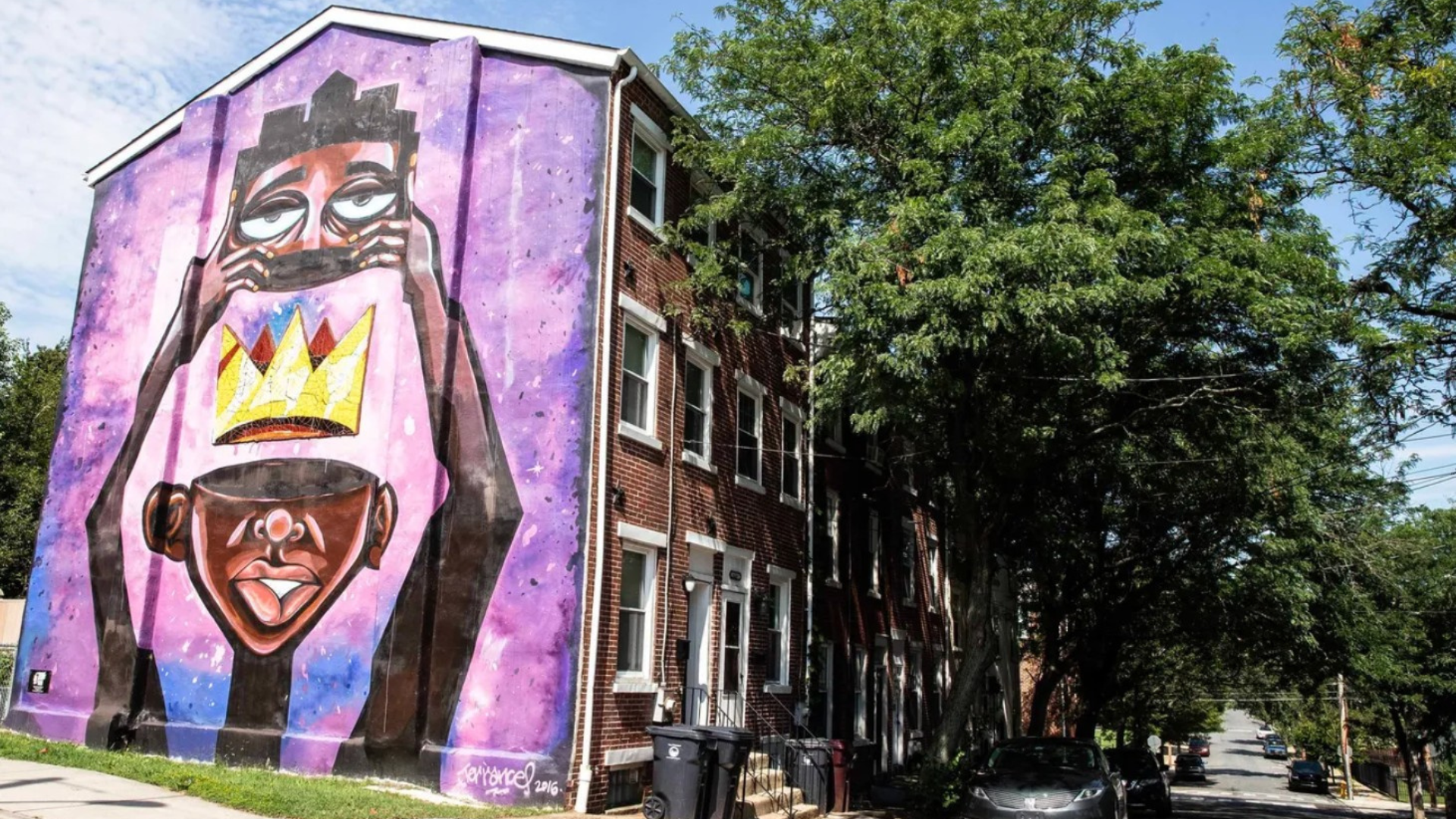 Large mural painted on the side wall of a brick rowhouse building, showing a stylized Black figure holding up a face or mask with a golden crown between two faces. The background is painted in purple and pink tones, and the mural rises above a tree-lined residential street.