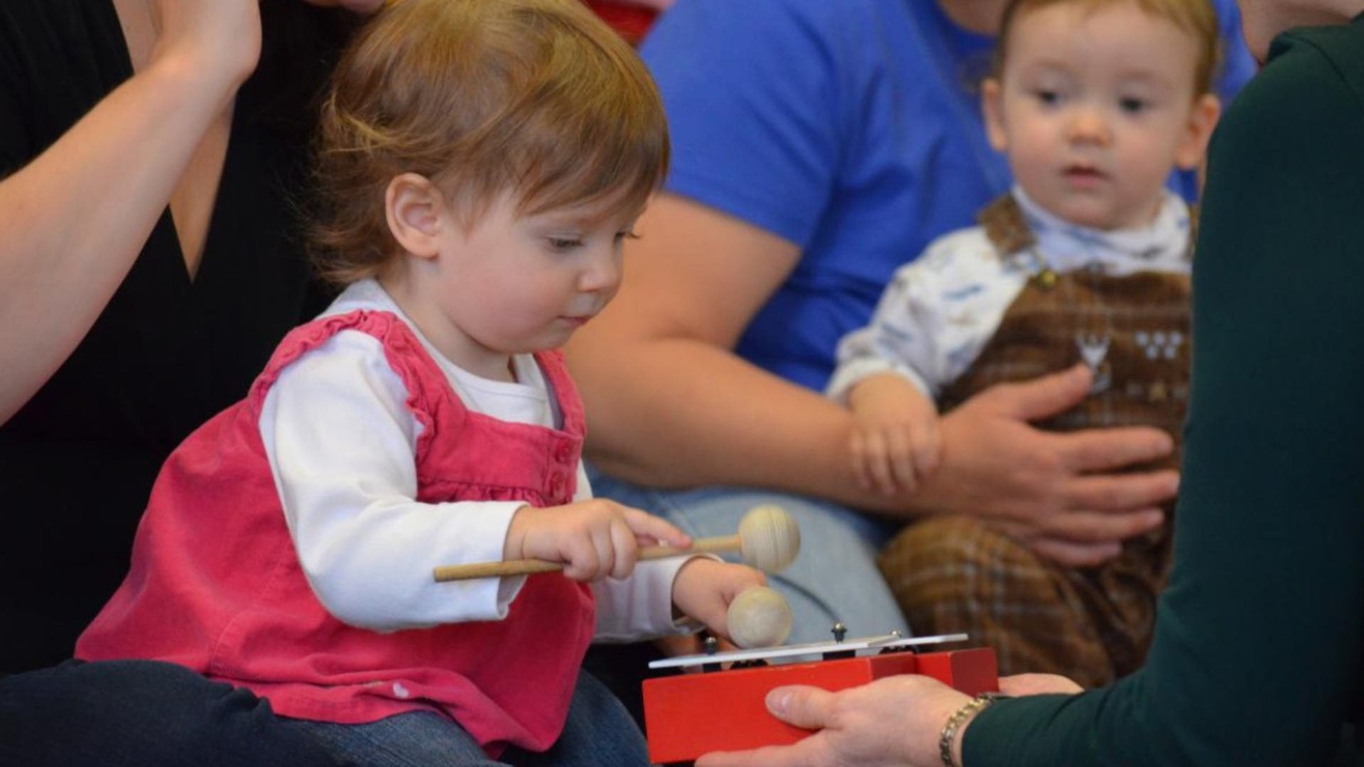 Young child seated among adults, concentrating while playing a small red percussion instrument with two mallets. Another toddler sits nearby on an adult’s lap, watching the activity.