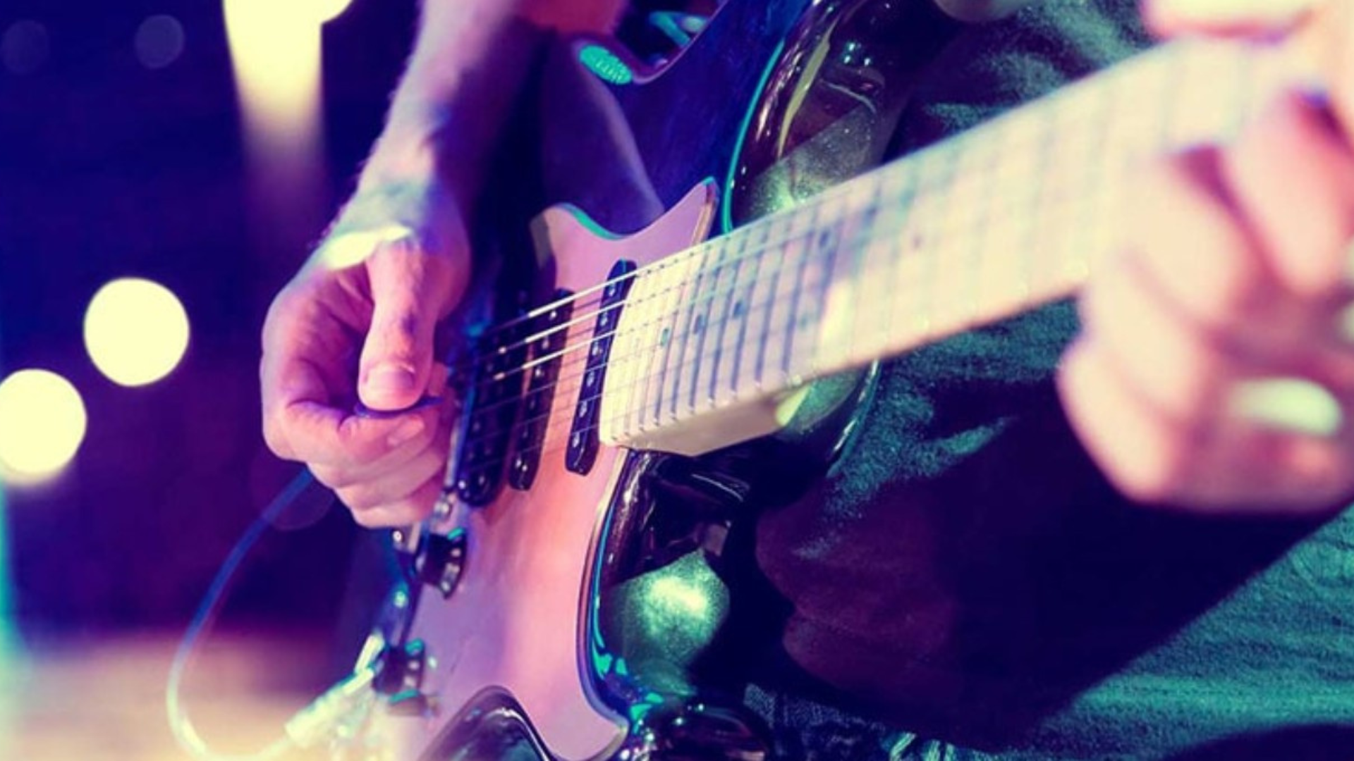 Close-up of a person playing an electric guitar under colorful stage lighting. The image focuses on the musician’s hands, strings, and pickups, with purple and white lights blurred in the background.