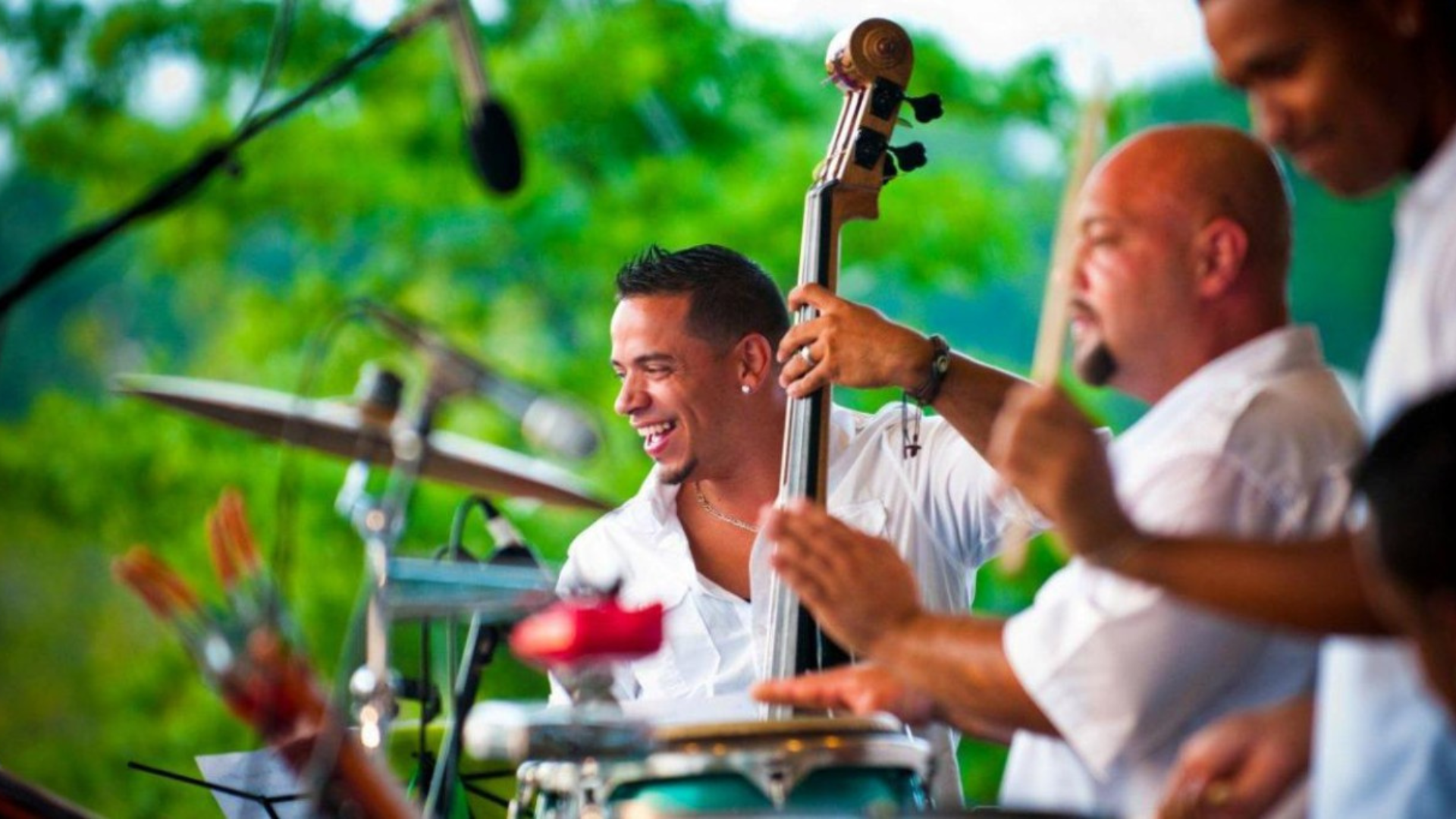 Musicians performing outdoors, with a smiling upright bass player in a white shirt at center and percussionists blurred in the foreground and background. Microphones and drums frame the scene, while lush green trees create a vibrant backdrop.