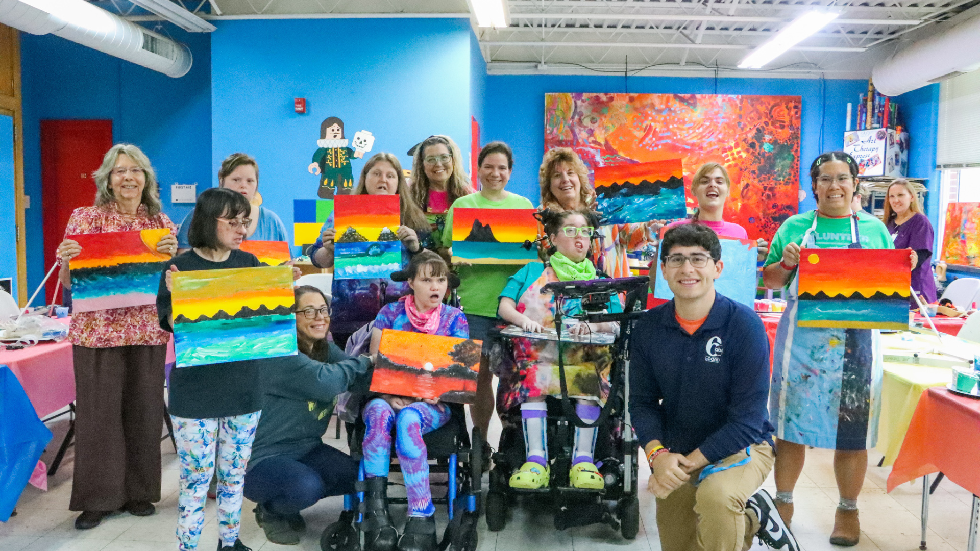 Group photo inside a bright art studio with blue walls, showing participants and volunteers holding colorful sunset and seascape paintings they created. Several people use wheelchairs, and tables with art supplies surround the group, creating a cheerful, inclusive workshop atmosphere.