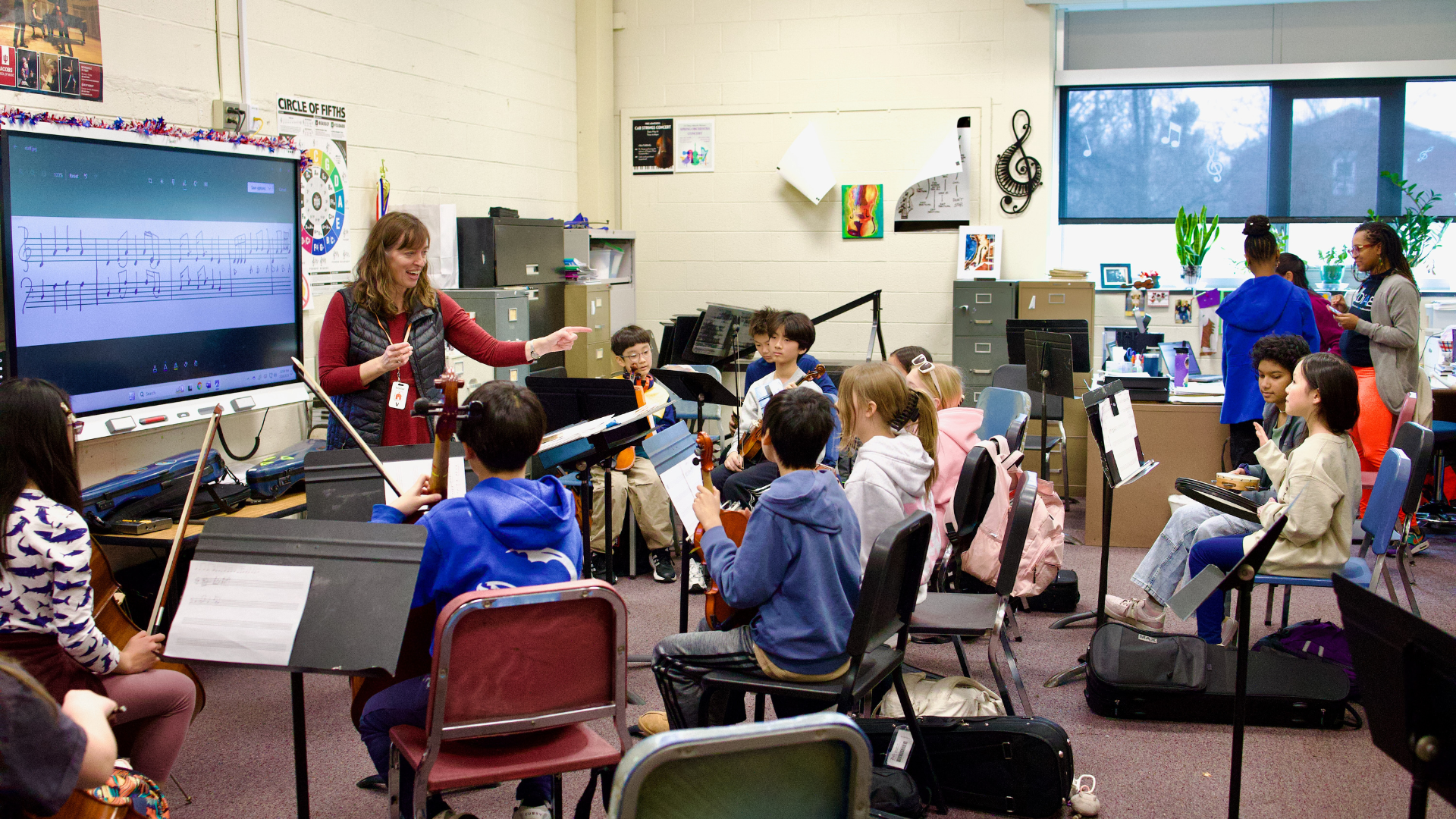Music teacher leading a classroom of students seated with string instruments and music stands during rehearsal. A large digital screen displays sheet music at the front of the room, while other students and staff work in the background of the bright, busy classroom
