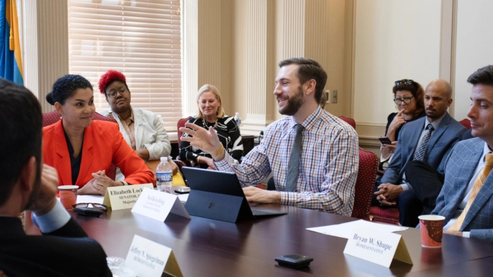 Group of officials and staff seated around a conference table during a formal meeting, with one man speaking and gesturing while others listen. Name placards, laptops, water bottles, and coffee cups are arranged on the table in a government-style meeting room with tall windows and paneled walls.