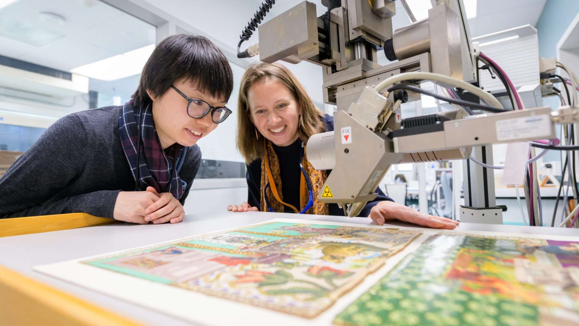 Two people lean over a large flatbed printer or cutting machine, closely examining a colorful printed artwork as the equipment hovers above the surface. The bright studio setting and their focused expressions suggest hands-on work in a printmaking or fabrication space.