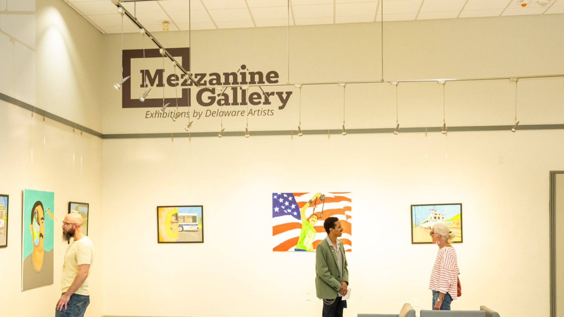 Wide view of the Mezzanine Gallery, a bright exhibition space with framed artworks hung along white walls beneath the sign “Mezzanine Gallery: Exhibitions by Delaware Artists.” Three visitors stand and walk through the gallery looking at the art, while a small seating area sits in the center of the room.