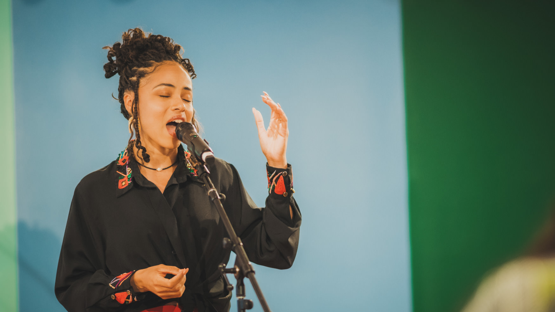 Singer performing into a microphone with eyes closed and one hand raised expressively. She wears a black blouse with colorful embroidered details, and the softly lit blue-green background keeps the focus on her face and gesture.