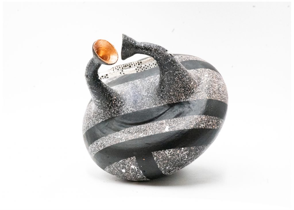 Round ceramic vessel with a textured black-and-silver surface decorated by broad dark bands. Two curved horn-like spouts rise from the top, one with a warm copper-colored interior, and a strip of reflective mosaic detail bridges the opening.