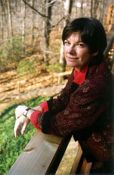 Portrait of a woman leaning on a wooden railing outdoors, wearing a dark textured jacket over a red blouse. She looks toward the camera with a gentle smile, while trees and a sunlit wooded landscape fill the softly focused background.