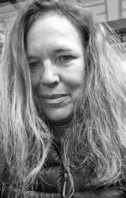 Black-and-white close-up portrait of a woman with long wavy hair, looking directly at the camera with a calm, slightly serious expression. The background is softly blurred, keeping the focus on her face.