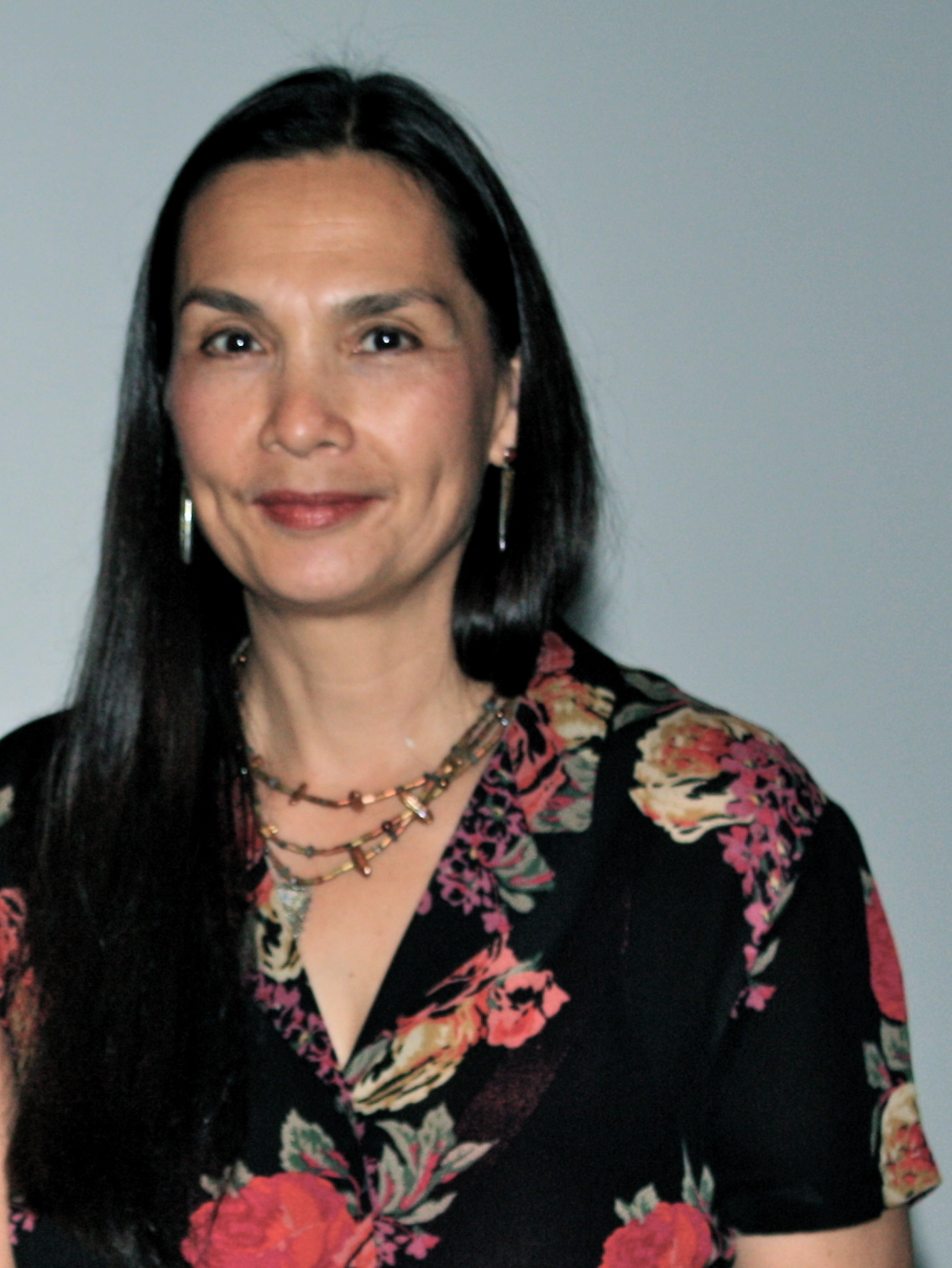 Portrait of a woman with long straight dark hair, wearing hoop earrings, layered necklaces, and a black floral top. She faces the camera with a slight smile against a plain light gray background.
