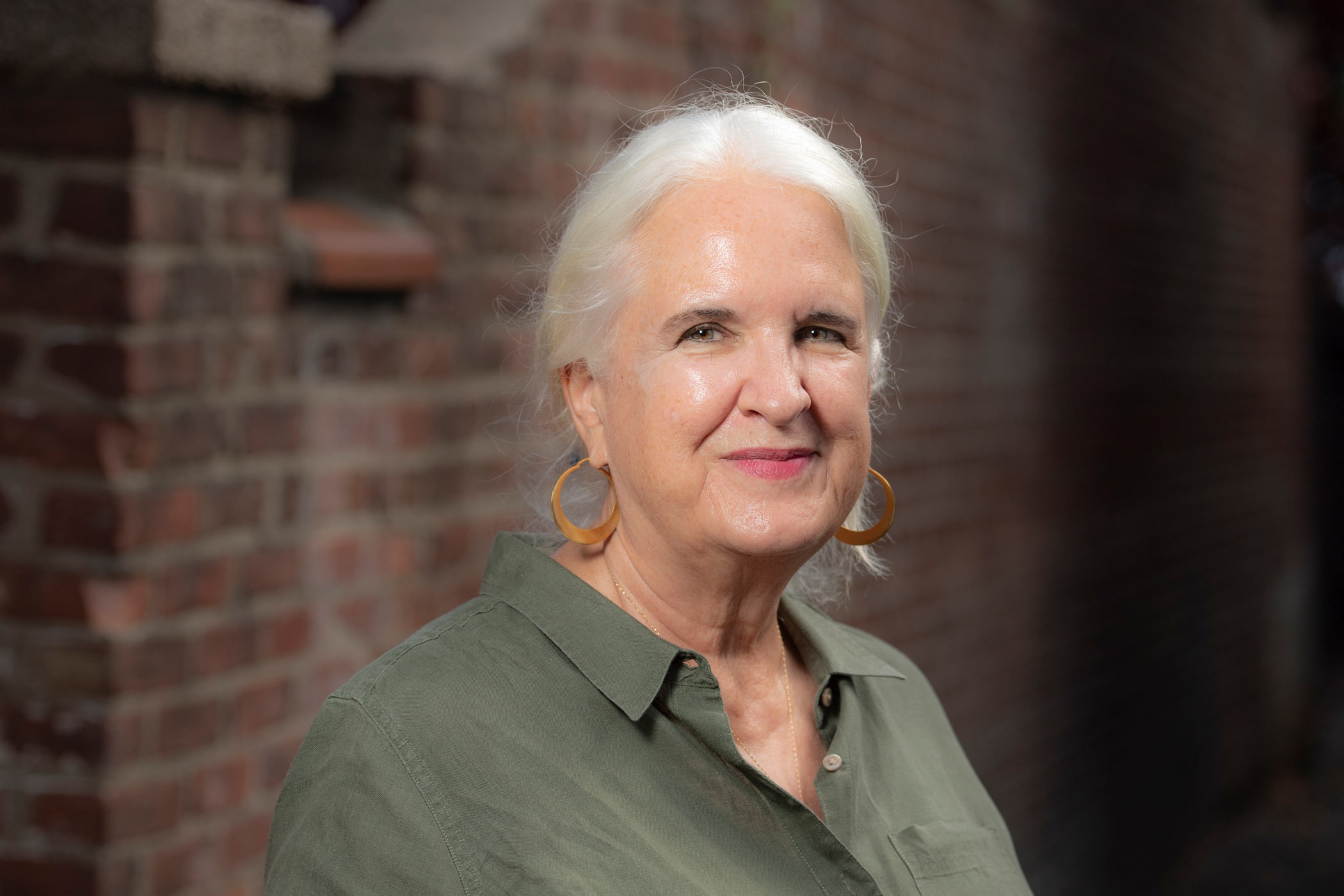 Portrait of a woman with white hair pulled back, wearing large hoop earrings and an olive green button-up shirt. She stands in front of a softly blurred brick wall, looking toward the camera with a calm, confident expression.