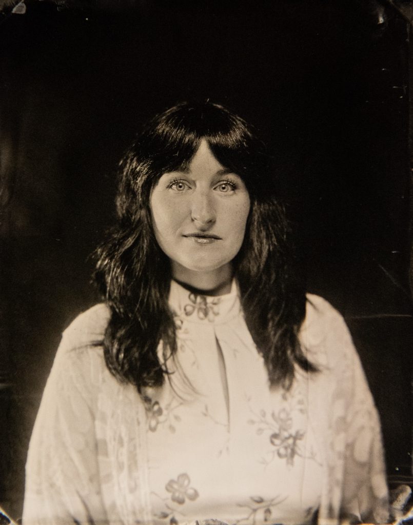 Black-and-white portrait of a woman with dark shoulder-length hair and bangs, wearing a light floral dress and a sheer shawl. She faces forward against a dark background, and the lighting emphasizes her eyes and the delicate details of her clothing.