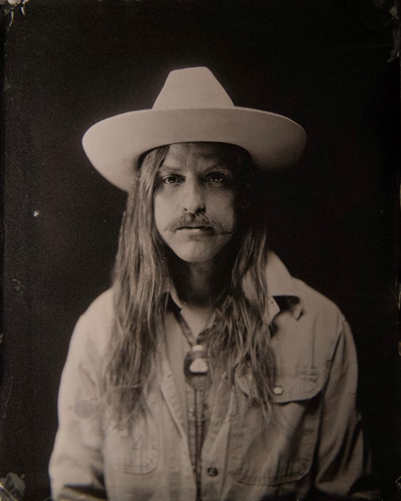 Black-and-white portrait of a person with long hair and a mustache wearing a wide-brimmed light-colored hat and a buttoned shirt. The subject faces the camera directly against a dark backdrop, giving the image a calm, vintage studio-portrait feel.