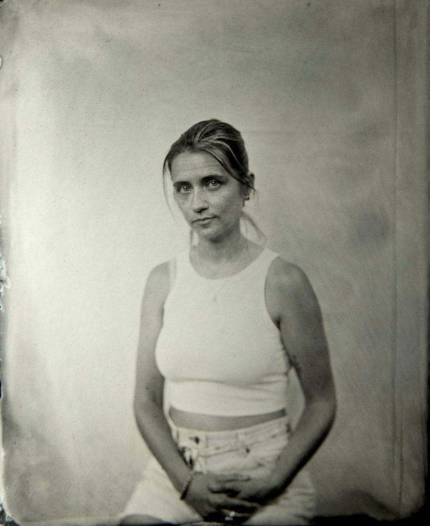 Soft-focus black-and-white portrait of a seated woman with her hair pulled back, wearing a sleeveless white top and light-colored bottoms. She sits with her hands folded in her lap against a plain backdrop, looking toward the camera with a gentle, reserved expression.