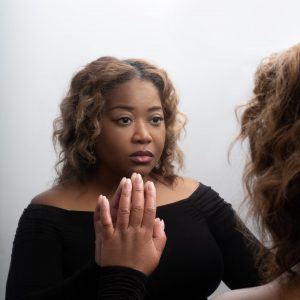 Portrait of a woman in a black off-the-shoulder top gazing thoughtfully at her reflection, with one hand raised against a mirror. The softly lit composition and neutral background create an intimate, reflective mood.