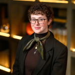 Portrait of a smiling person with short curly hair, glasses, and a patterned neck scarf, wearing a dark blazer and standing indoors before warmly lit shelves.