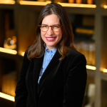 Portrait of a smiling woman with glasses, shoulder-length brown hair, and a black blazer over a light blue ruffled blouse, standing indoors before warmly lit shelves.
