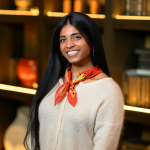 Portrait of a smiling woman with long black hair, hoop earrings, and a bright patterned scarf, standing indoors before warmly lit shelves with a softly blurred background.