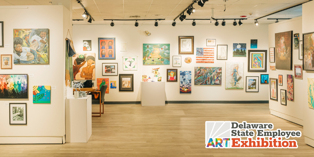 Wide view of a gallery filled with colorful framed artwork hung salon-style across white walls, with track lighting overhead and a light wood floor. The “Delaware State Employee ART Exhibition” logo appears in the bottom right corner.
