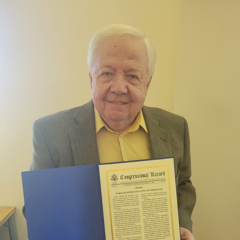 A man in a yellow button up and brown blazer smiles at the camera while holding a proclamation