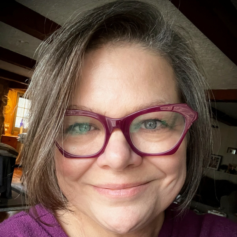 A woman smiles into the camera. She has brown hair and purples glasses
