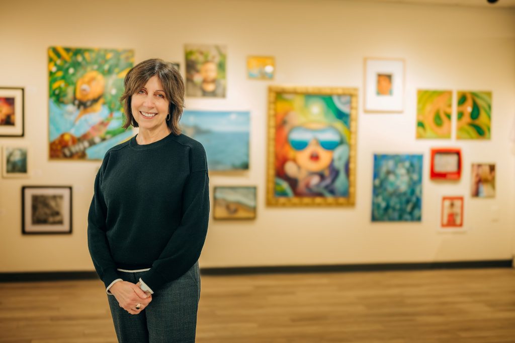 A smiling woman stands in an art gallery with her hands clasped in front of her, wearing a dark sweater and gray pants. Behind her, a wall of colorful framed artworks is softly blurred, creating a warm and welcoming portrait in an exhibition space.