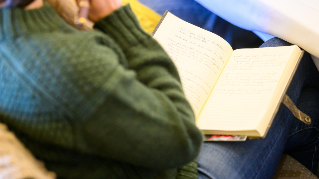 Close-up of a person in a green sweater writing notes in an open notebook resting on their lap. The page is filled with handwritten reflections, suggesting a quiet moment of writing, journaling, or workshop participation.