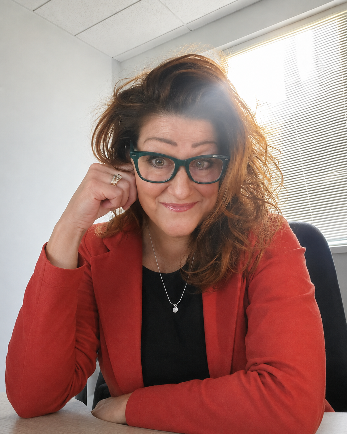 Casual headshot of a smiling woman with long reddish-brown hair and dark-framed glasses, resting her head lightly on one hand. She wears a red jacket over a black top and is seated indoors near a bright window with blinds.
