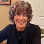 A white woman with short hair, wearing glasses and a black sweater, sitting at a table inside.