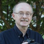 A bald white man in glasses, smiling, outside in front of a tree.