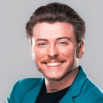 A white man with brown hair and a mustache, wearing a teal suit jacket in front of a gray wall.