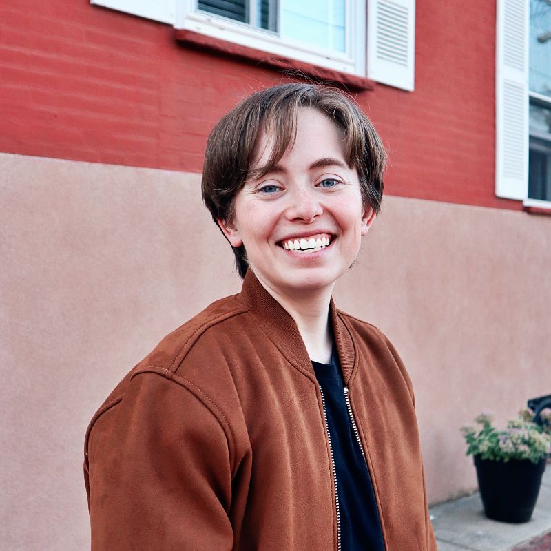 A young white person with a short haircut, smiling, outside in front of a red wall.