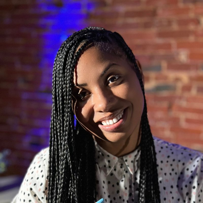 A black woman with braids in a short-sleeve button-up shirt, holding up a peace sign. She is indoors in front of a brick wall.