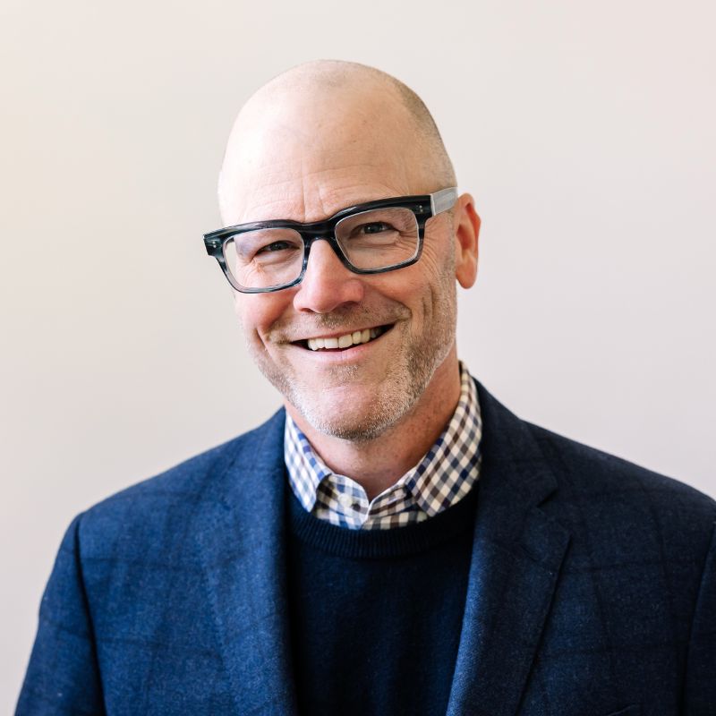 A bald white man in glasses and a blue suit jacket, in front of a white wall.