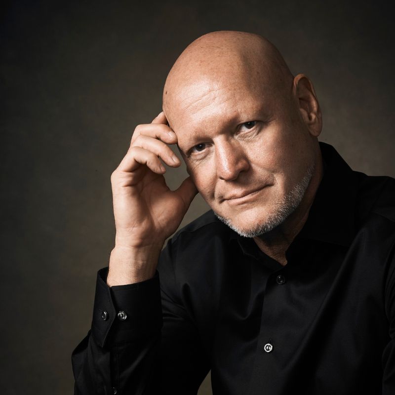 A headshot of a bald white man in a black button up shirt.