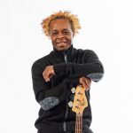 A black man with blonde curly hair, in a black jacket, smiling and crossing his arms. Ibn front of a a white background.