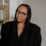 A headshot of a black woman with long box braids, wearing glasses and a large black suit jacket. She is in front of a white wall with a gold frame.