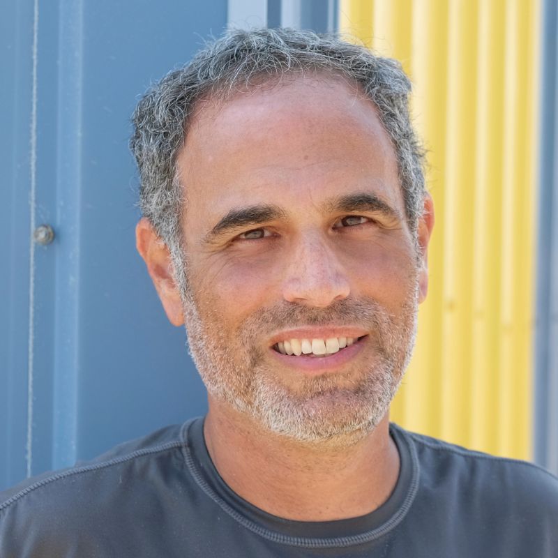 A man with gray hair in a gray shirt, in front of a blue and yellow background.